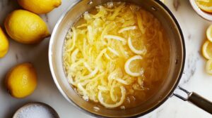 Citron slices simmering in a pot of syrup, surrounded by fresh lemons and a bowl of sugar