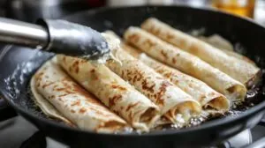 Close-up of Xango rolls being fried in a skillet, with golden brown tortillas sizzling in oil, held by tongs