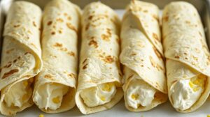 Close-up of four tortillas filled with creamy white filling and bits of corn, neatly rolled and arranged in a row