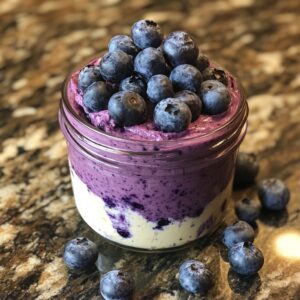 A jar of blueberry cheesecake topped with fresh blueberries on a marble countertop, with scattered blueberries around