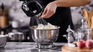 Person using a black stand mixer to whip marshmallow mixture in a stainless steel bowl on a marble countertop