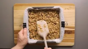 A Person Using a Spatula to Press the Protein Bar Mixture Evenly Into a Parchment-Lined Baking Dish