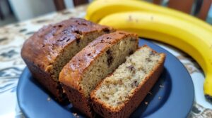 A freshly baked loaf of banana bread on a plate, partially sliced, with ripe bananas in the background