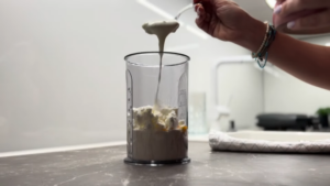 A Person Adds a Spoonful of Blended Batter Into a Tall Measuring Cup with Cottage Cheese, Eggs, and Oats on A Kitchen Counter