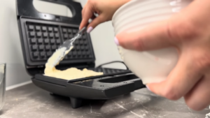 A Person Spreads Waffle Batter onto A Preheated Waffle Iron Using a Spatula While Holding a White Mixing Bowl