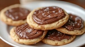 A plate of golden, soft-baked cookies topped with glossy Nutella drizzle and a touch of sea salt