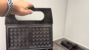 A Person Lifts the Lid of A Black Waffle Iron on A Kitchen Counter, Preparing to Preheat It